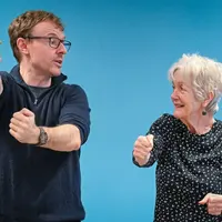 A man demonstrates a martial arts movement standing next to an older woman. The woman is copying the movement.