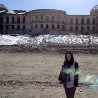 A woman wearing a headscarf stands in front of large, damaged building. There is snow on the ground in front of the building.