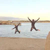 Two people wearing wetsuits jump into the water from a concrete dock