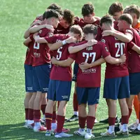 A group of young men huddled together on a football pitch. They are wearing maroon shirts and dark blue shorts.