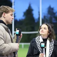 Two people stand on a rugby pitch. They are holding microphones with Met TV printed on them.