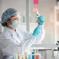 A woman in white lab coat, wearing safety equipment, analyses a red liquid in a test tube