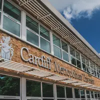 A building with large windows featuring the sign Cardiff Metropolitan University on a brick wall. A blue sky with clouds is visible above.