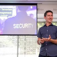 A man stands in front of a screen giving a presentation. The screen shows a person wearing a shirt with SECURITY printed on the back.