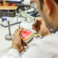 A dental technician works on a dental mold, meticulously shaping it with tools in a well-equipped lab. A small flame burner is visible on the table, suggesting a process that involves heat.