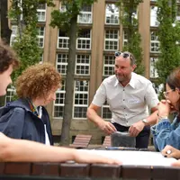 Four people sit at a wooden table outdoors. A man wearing a name badge labelled Gary stands next to them.