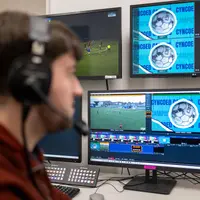 A person in headphones watches a football match on several monitors.