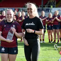 A female rugby player poses with an award in front of her teammates and alongside another individual