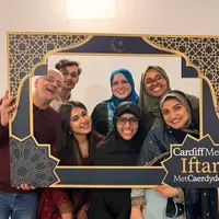 A group of people pose for a photo holding a decorative frame. In the corner of the frame is written Cardiff Met Iftar.