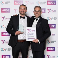 Two men in suits proudly hold an award in front of a decorative wall.  =
