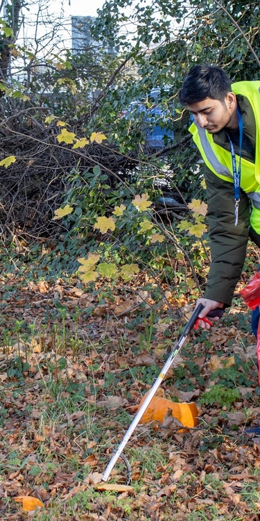 Young adult in high visibility jacket uses a litter picker to collect rubbish in a large red bin bag