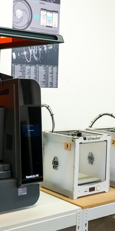 A collection of 3D Printers lined up on a row of desks