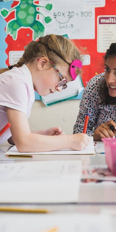 A school teacher sits at a table next to two young children. The children are writing in notebooks.