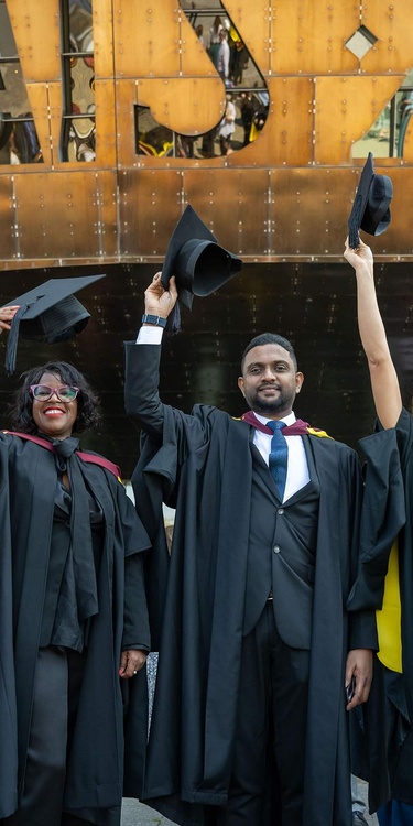 Six students in graduation gowns line up in front of the Wales Millennium Centre, holding their caps in the air.