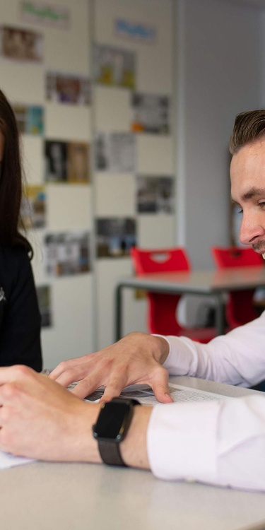 A teacher kneels next to a pupil sitting at a desk, pointing to the open page of a book.