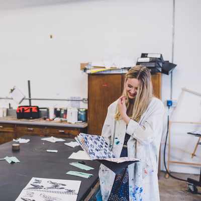 Three women wearing paint-splattered lab coats work together in a classroom. One woman, in a green shirt, smiles and looks down. The other two women, one wearing a hijab, also smile while focusing on what theyre doing. Art supplies are visible.