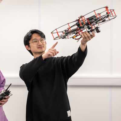 A person with long hair is sitting at a table with a laptop, interacting with a humanoid robot. They appear to be adjusting or programming the robot. The setting is a modern, brightly lit room with yellow chairs.
