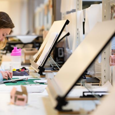 A person wearing a black shirt and black pants is focused on woodworking, marking a piece of wood with a pencil. Safety glasses rest on the table. In the background, a window and machinery are visible in a bright workshop.