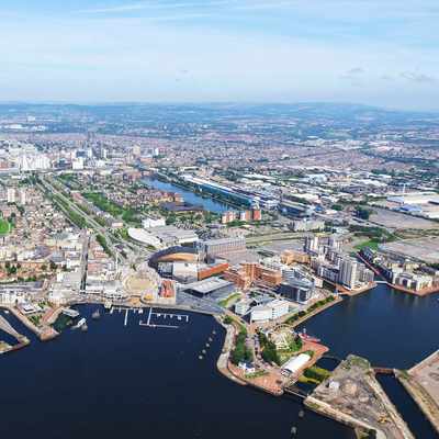 Aerial view of a vibrant waterfront city with winding rivers, numerous buildings, and a marina. The landscape features green spaces and urban areas under a clear blue sky.