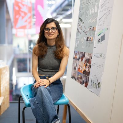 A young woman with long brown hair smiles while sat on a stool. Beside her are images and designs pinned to a whiteboard.