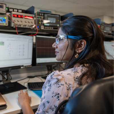 A person with blond hair, wearing a blue patterned shirt, is focused on working with a small robot or electronic device using a screwdriver at a desk in a lab or office setting. Various electronic equipment is visible in the background.