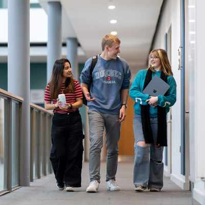 Three young people walk together on a balcony. One is holding a laptop computer.