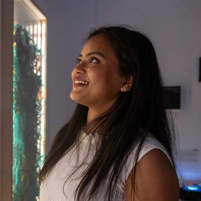 A woman with long dark hair and a white sleeveless top smiles while looking at a brightly lit art exhibit featuring various colorful pieces mounted on a wall. The room is dimly lit, enhancing the glowing display.