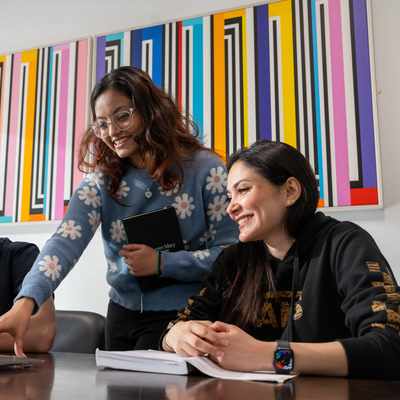 Three people are gathered around a laptop at a table. One person is pointing at the screen, while the others smile and look on. A colorful abstract painting is on the wall behind them.
