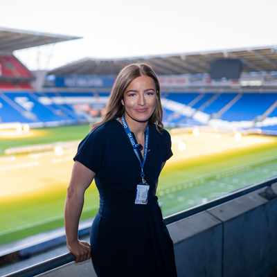 A woman in a navy dress stands smiling on the balcony of an empty stadium. The stadium seats are predominantly blue, with some red sections, and green grass covers the field. She wears an ID lanyard around her neck.