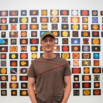 A person wearing a cap stands in front of a large wall collage featuring numerous colorful images of celestial bodies, like stars and planets. The collage is composed of small, square pictures arranged in a grid pattern.