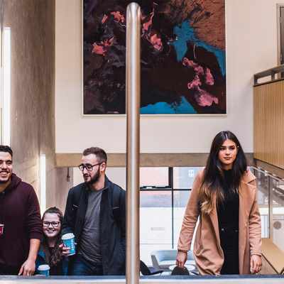 Four people are walking up a staircase, carrying coffee cups. They appear to be engaged in conversation and smiling. The setting is modern with concrete walls and a colorful abstract painting on the upper wall.