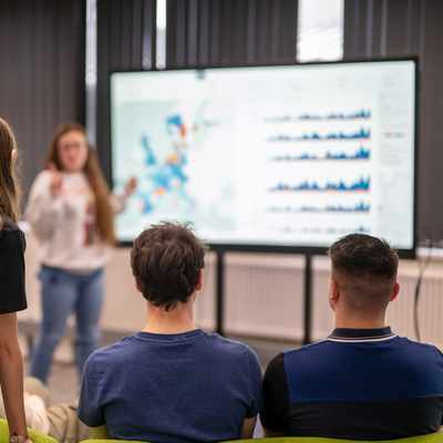 Four people are seated and watching a presentation on data displayed on a large screen, while one person stands in front of the screen explaining the content. The room has a professional setting with green chairs.