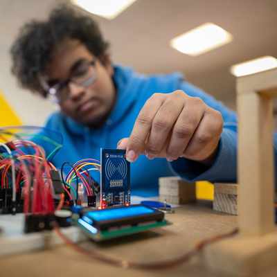 A person in a blue hoodie works with electronic components and circuit boards, placing a small blue board among connecting wires on a table. The background is blurred and the lighting is bright.