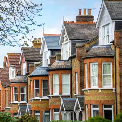 A row of traditional two-story brick houses with bay windows and gabled roofs in an urban neighborhood. Leafy trees line the street, and the sky is partly cloudy, adding a serene backdrop to the scene.