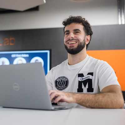 A person with a beard is smiling while using a laptop in a modern workspace. In the background, theres a screen and a wall with The Learning Lab text on it. They are wearing a white shirt with patches.