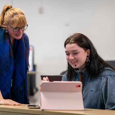 A teacher with glasses and a blue scarf leans over a students desk in a classroom. The student, smiling, is using a pink tablet. Other students appear in the blurred background.