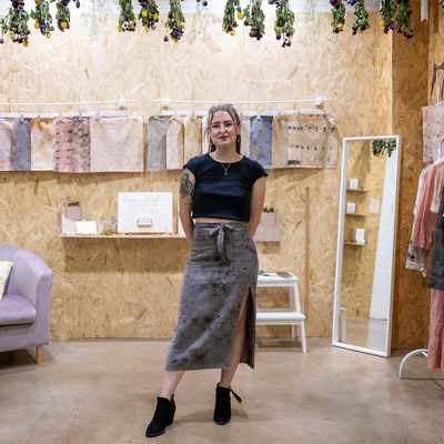 A woman stands smiling in a cozy, wood-paneled boutique. She wears a black top and a long skirt. The room features dried flowers, a chair with a patterned pillow, art on the walls, and display shelves with items.