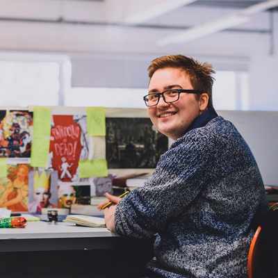 A person with short hair and glasses smiles while sitting at a desk covered in colorful images and sticky notes. They hold a pen and notebook, looking towards the camera in a bright, open office space.