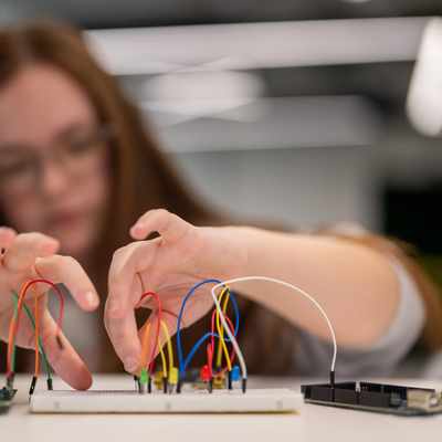 A person with long hair and glasses is working with a breadboard and wires, connecting electronic components. The background is blurred, focusing on the hands and the breadboard.