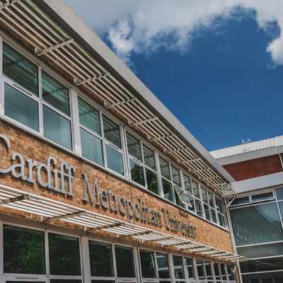 The image shows the exterior of Cardiff Metropolitan University. The building has a modern design with large windows and the universitys name prominently displayed in white lettering on a brick facade under a blue sky.