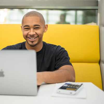 A person is smiling while sitting at a table with a laptop in front of them. They are in a bright, modern setting, with a yellow cushioned seating area behind them.