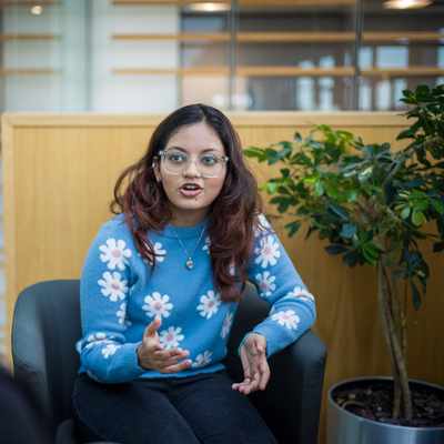 A young woman with glasses and long hair, wearing a blue sweater with white flower patterns, gestures while talking. She is seated in a modern indoor space, facing another person whose back is to the camera. A green plant is in the background.