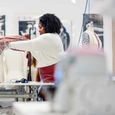 A person with curly hair holds up a colourful fabric in a design studio. In the background, a mannequin and framed art are visible. The room is well-lit, creating an inspiring workspace for creativity.