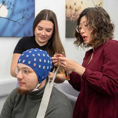 Two people assist a seated person with an EEG cap. One adjusts the cables on the cap, while the other looks on.