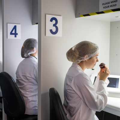 A row of people in white lab coats and hairnets sit at numbered stations. Each person is engaging in a sensory evaluation, with one person smelling or tasting from a small cake under a hooded light. The stations are numbered 3 to 8.