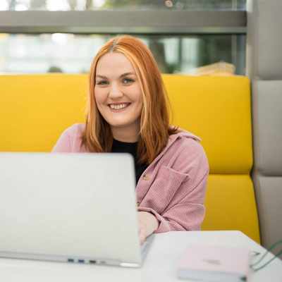 A young woman is smiling while sitting at a table with a laptop in front of them. They are in a bright, modern setting, with a yellow cushioned seating area behind her.