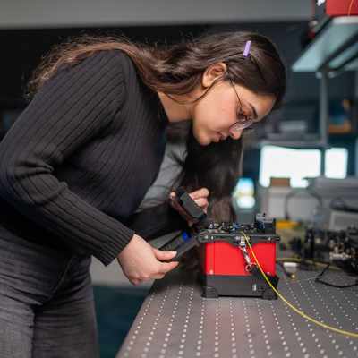 A student pressing the touchscreen of a small electronic device. In the background are other devices and computer monitors.