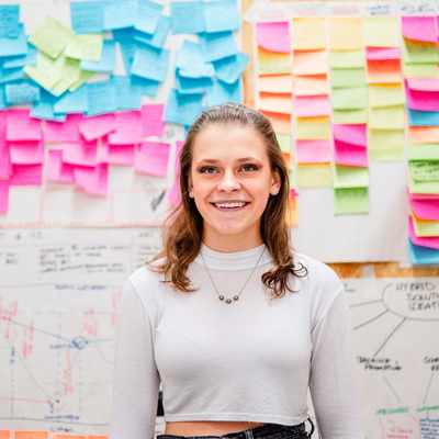 A woman stands smiling in front of a wall covered with colourful sticky notes and charts. The notes are pink, blue, green, and yellow, suggesting a brainstorming or planning session. The background includes diagrams and handwritten notes.