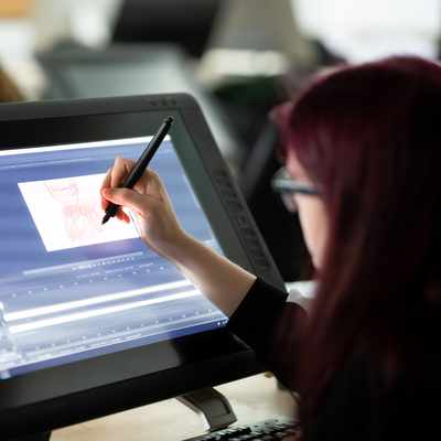 A person with long hair is using a stylus to draw on a large digital tablet. The screen displays graphic design software with artwork in progress. Other people are visible in the background, working at similar stations.