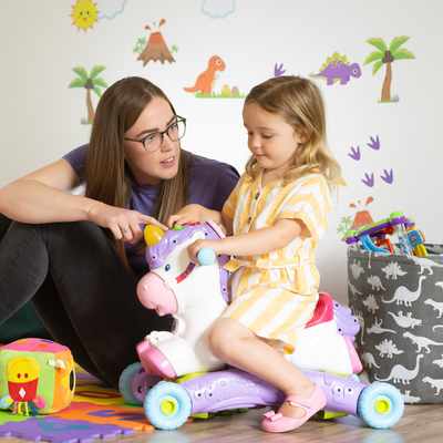 A woman with glasses plays with a young girl on a unicorn ride-on toy. They are surrounded by colorful toys and decorations, including a xylophone cart and building blocks, in a room with playful wall stickers.
